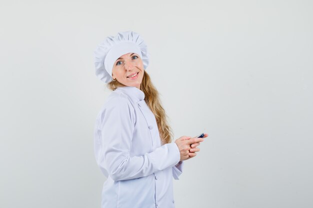 Female chef posing while holding cellphone in white uniform and looking merry .
