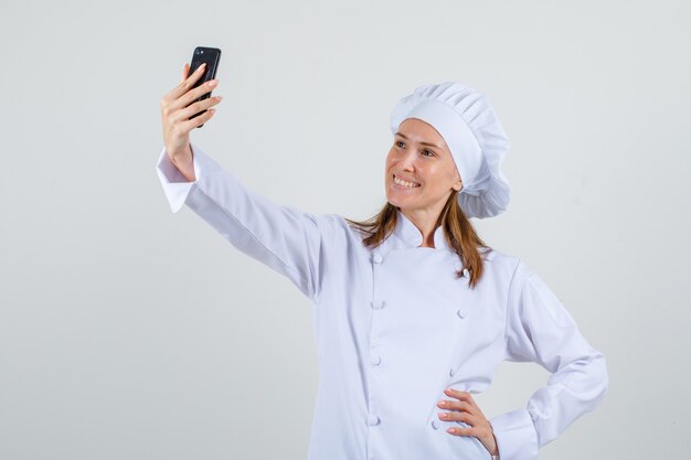 Female chef posing while taking selfie in white uniform and looking cheerful