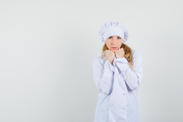 Female chef propping chin raised fists in white uniform and looking focused 