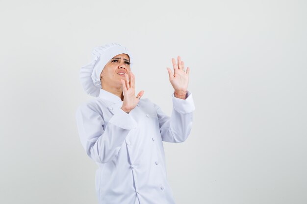 Female chef raising palms to defend herself in white uniform and looking scared. 