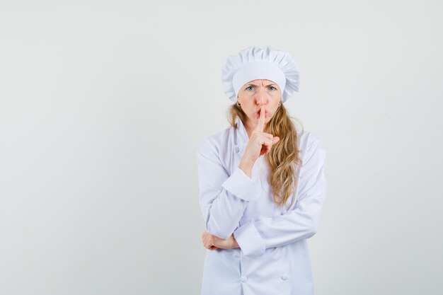 Female chef showing silence gesture while frowning in white uniform and looking serious. 