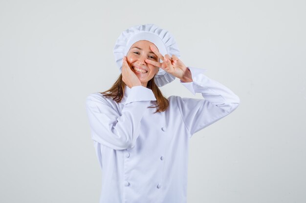 흰색 유니폼에 눈 근처 v 기호를 표시 하 고 쾌활 한 찾고 여성 요리사. 전면보기.