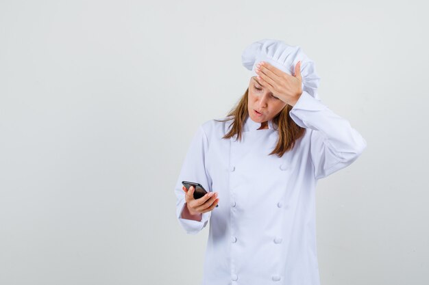 Female chef using smartphone with hand on forehead in white uniform and looking regretful. front view.