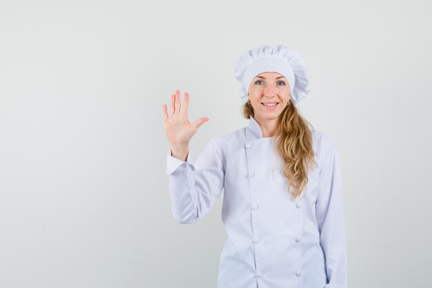 Female chef waving hand to say hello or goodbye in white uniform and looking cheerful 