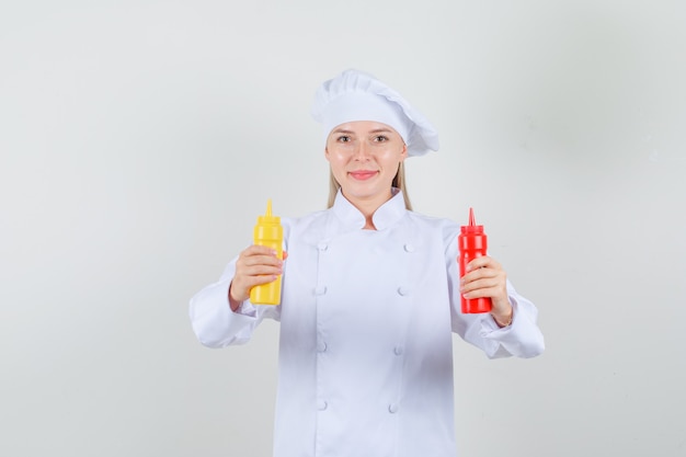 Female chef in white uniform holding bottles of ketchup and mustard and looking cheerful 