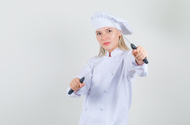 Female chef in white uniform holding knives in boxer pose and looking glad 