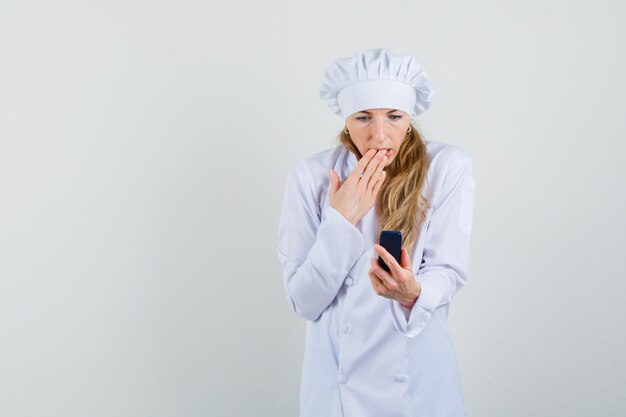 Женщина-повар в белой форме смотрит на мобильный телефон и выглядит удивленно