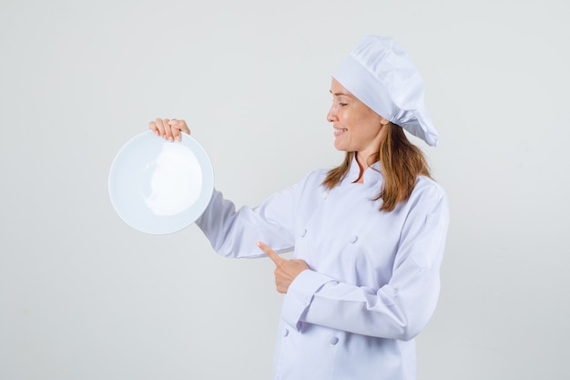 Female chef in white uniform pointing finger at empty plate and looking glad