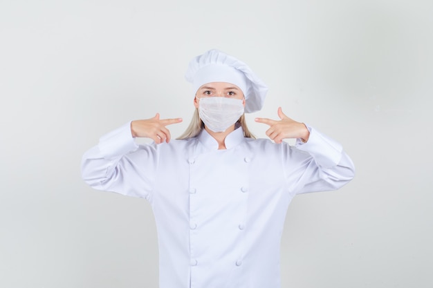 Женщина-повар в белой форме, указывая пальцами на медицинскую маску