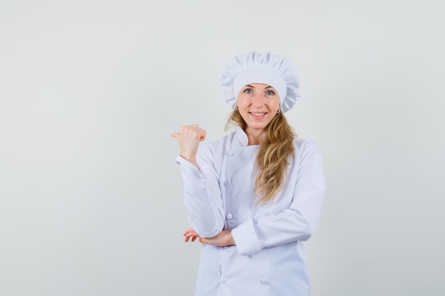 Female chef in white uniform pointing thumb up back and looking merry 