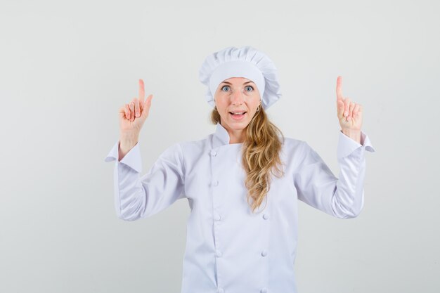 Женщина-повар в белой униформе указывает вверх и выглядит весело