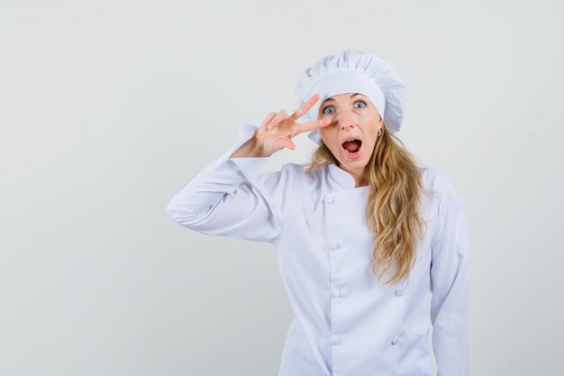 Женщина-шеф-повар в белой форме показывает знак v возле глаза и выглядит сумасшедшим