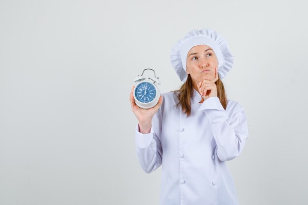 Female chef in white uniform thinking while holding alarm clock and looking hopeful