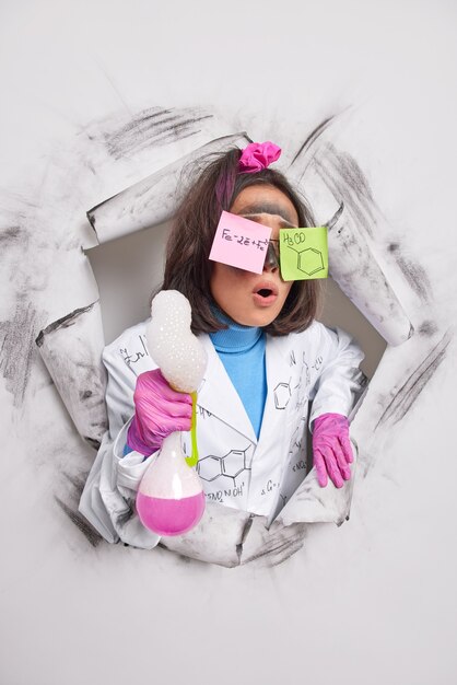 female chemist has stuck stickers on eyes with written formulas holds flask with pink liquid bubbles wears white coat rubber gloves keeps mouth opened from wonder breaks through paper hole