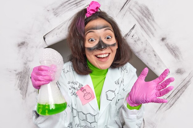 female chemist holds beaker with mixed reagents conducts scientific research wears white coat with stuck sticker drawn formulas rubber gloves smiles pleasantly breaks through paper 