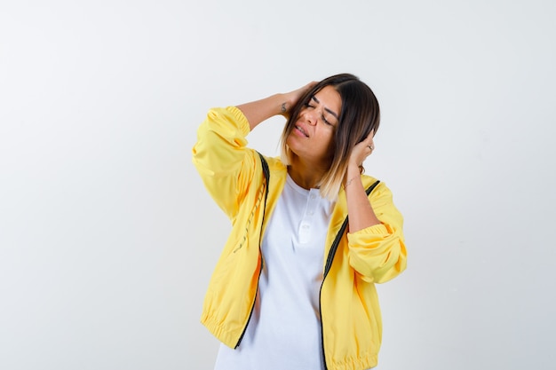 Female clasping head in hands in t-shirt, jacket and looking relaxed , front view.