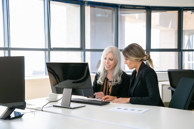 Free photo female colleagues sitting at workplace together, using computer near paper diagram. business communication or mentorship concept