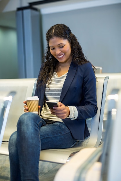 Pendolare femminile con tazza di caffè utilizzando il telefono cellulare in sala d'attesa