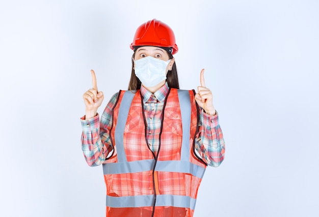 Female construction engineer in red helmet and safety mask pointing up.