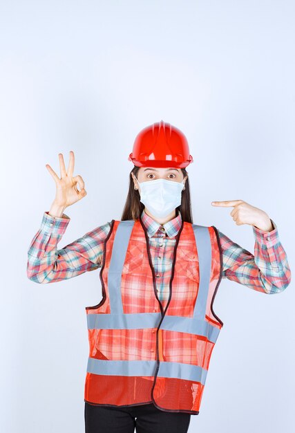 Female construction engineer in red helmet and safety mask showing successful hand sign