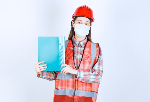 파란색 폴더를 들고 확인을 위해 제시하는 안전 마스크와 빨간색 헬멧을 쓴 여성 건설 엔지니어