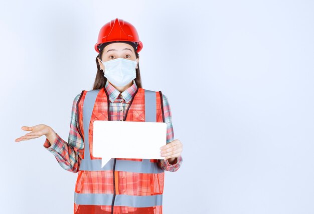 Female construction engineer in safety mask and red helmet holding a rectangle blank info board and looks confused and thoughtful