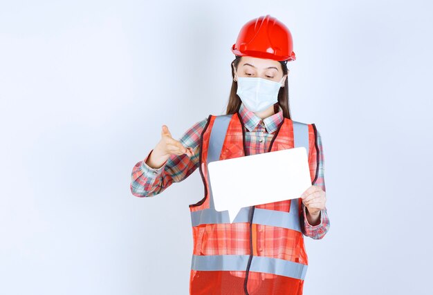 Female construction engineer in safety mask and red helmet holding a rectangle blank info board