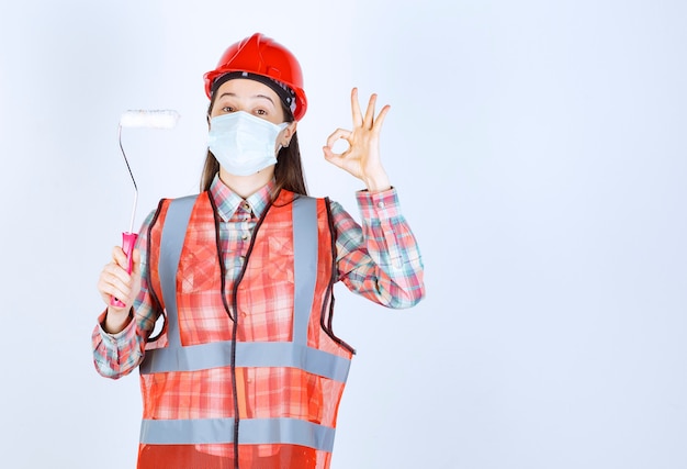 Female construction engineer in safety mask and red helmet holding a trim roller for painting and showing enjoyment sign