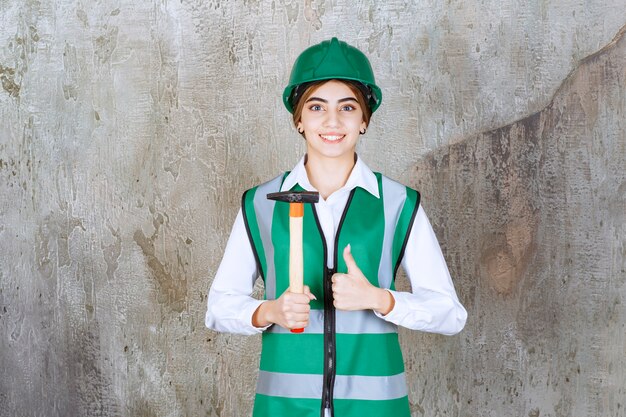Female construction worker in green helmet holding hammer and giving thumbs up