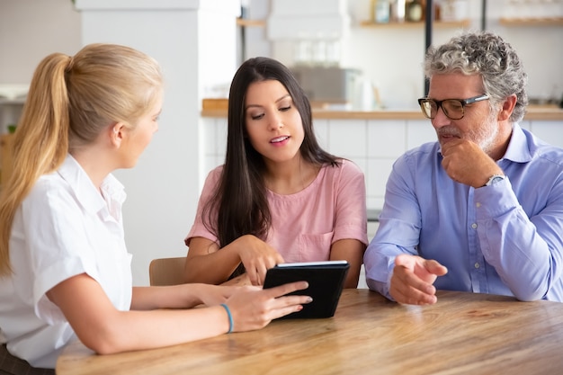 Female consultant or manager meeting with couple of young and mature customers, presenting content on tablet