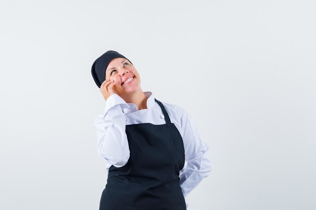 Cuoco femminile che tiene la mano sulla guancia in uniforme, grembiule e sembra sognante. vista frontale.