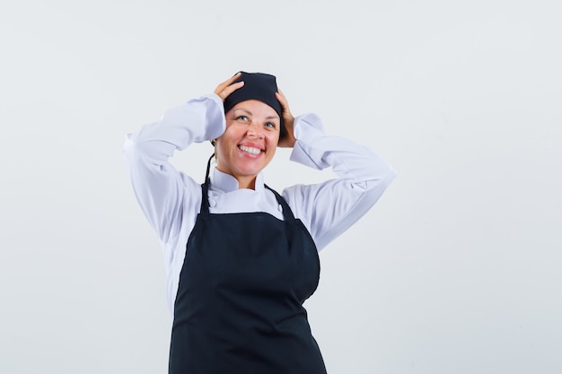 Female cook holding hands on head in uniform, apron and looking joyful , front view.