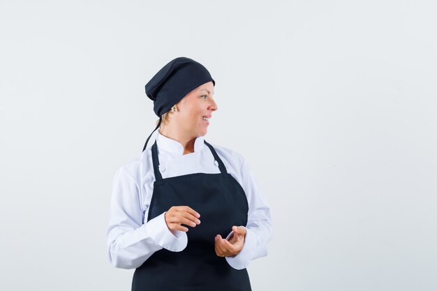 Female cook looking aside in uniform, apron and looking cheerful , front view.