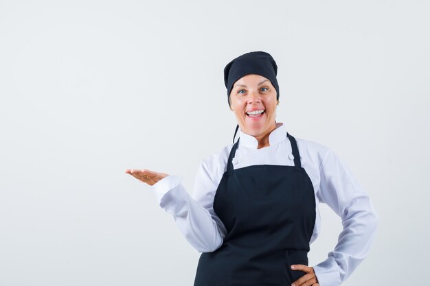 Female cook pretending to hold something in uniform, apron and looking glad. front view.