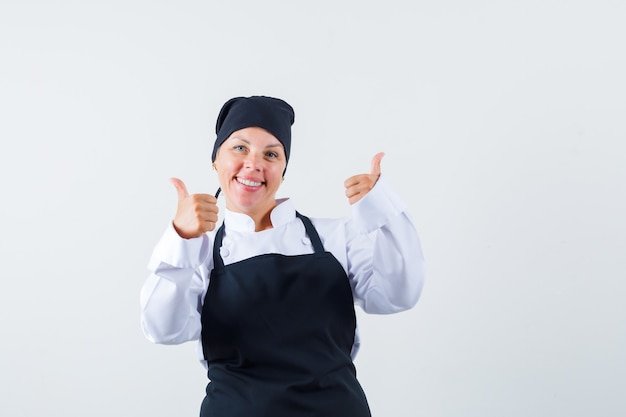 Female cook showing double thumbs up in uniform, apron and looking merry , front view.