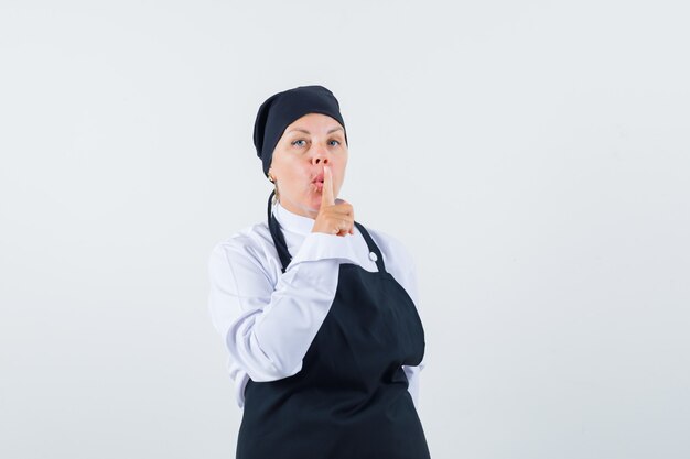 Female cook showing silence gesture in uniform, apron and looking careful. front view.