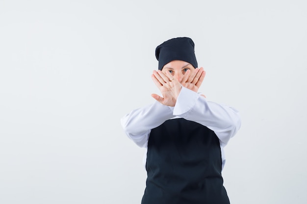 Женщина-повар показывает жест остановки в униформе, фартуке и выглядит серьезным, вид спереди.