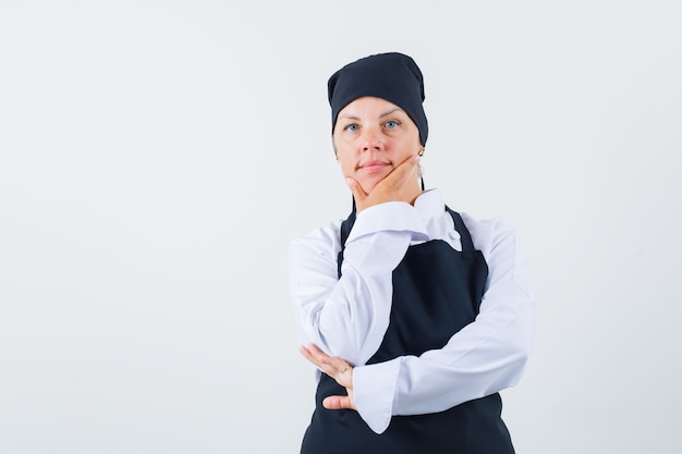 Female cook in uniform, apron holding hand on chin and looking sensible , front view.