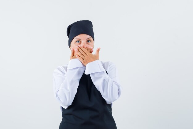 Female cook in uniform, apron holding hands on mouth and looking surprised , front view.