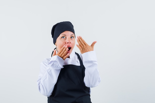 Cuoco femminile in uniforme, grembiule che tiene le mani vicino alla bocca aperta e che sembra sorpreso, vista frontale.