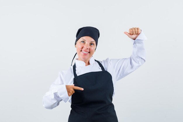 Female cook in uniform, apron pointing aside, pretending to hold something and looking cheerful , front view.