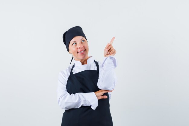 Female cook in uniform, apron pointing up and looking dreamy , front view.