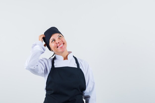 Женщина-повар в униформе, почесывает голову в фартуке и выглядит веселой, вид спереди.