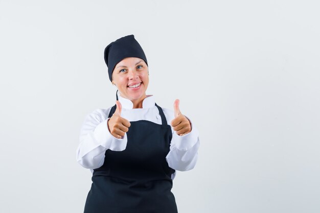 Female cook in uniform, apron showing double thumbs up and looking cheery , front view.