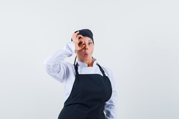 Female cook in uniform, apron showing ok sign on eye and looking amazed , front view.