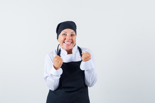 Female cook in uniform, apron showing winner gesture and looking happy , front view.