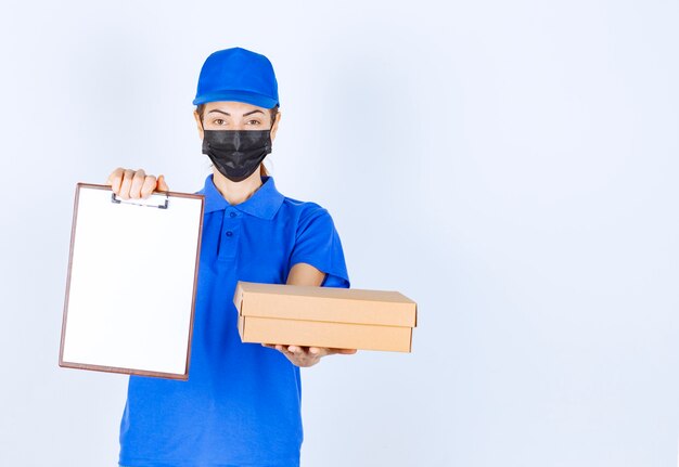 Female courier in blue uniform and face mask delivering a cardboard parcel and asking the customer to sign on the blank. 