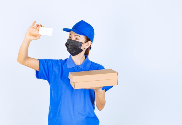 Female courier in blue uniform and face mask delivering a cardboard parcel and presenting her business card . 