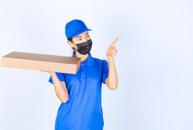 Corriere femminile in uniforme blu e maschera facciale che tiene una scatola di cartone e indica da qualche parte.