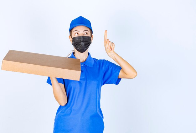파란색 유니폼과 얼굴 마스크를 쓴 여성 택배사는 판지 상자를 들고 어딘가를 가리키고 있습니다.